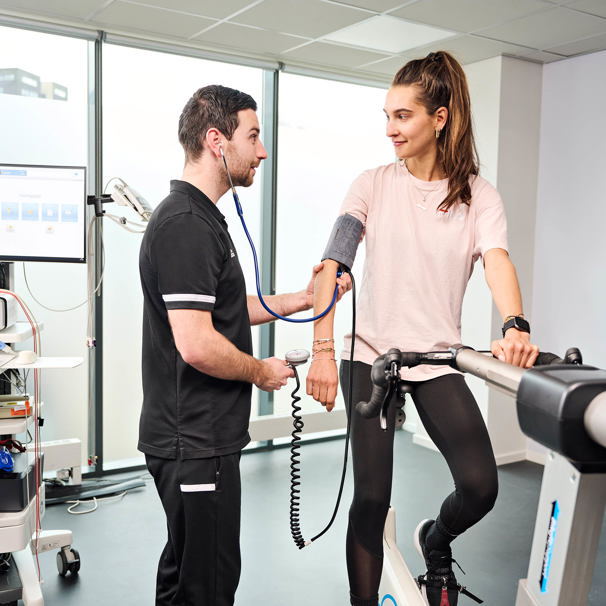 Een sporter zit op de hometrainer. De inspanningsfysioloog meet de bloeddruk van de sporter. Met een sportkeuring brengt Anna TopSupport jouw gezondheid en belastbaarheid in beeld. We hebben verschillende sportkeuringen of sportonderzoeken.