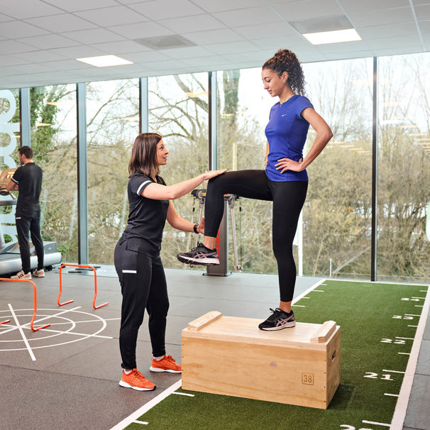 Een sporter staat in de oefenzaal op een houten blok. 1 knie is gebogen. De manueel therapeut houdt de knie vast en geeft uitleg. Als je ongemak hebt bij het bewegen, helpt Anna TopSupport je verder.