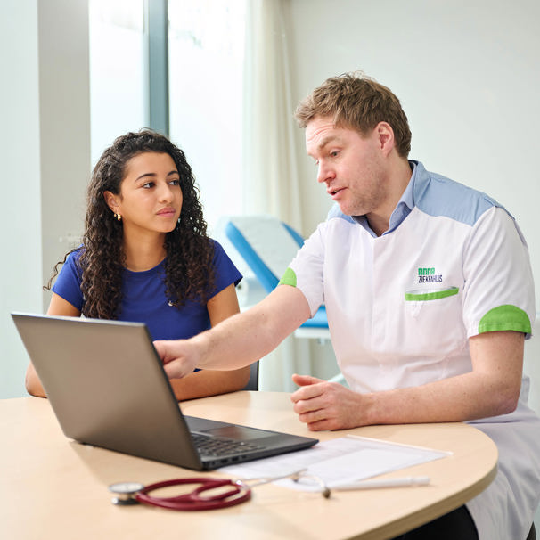 Een patiënt zit aan tafel met de dokter die uitleg geeft en iets aanwijst op een laptop. Het Basisplus Sport Medisch Onderzoek is geschikt als je zonder klachten sport en tussen de 12 en 35 jaar oud bent.