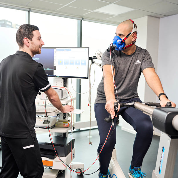 Een sporter zit op een hometrainer en draagt een ademmasker. De inspanningsfysioloog De inspanningsfysioloog staat bij de sporter en heeft een beeldscherm met meetapparaten. Het Uitgebreid Sport Medisch Onderzoek bestaat uit het Groot Sport Medisch Onderzoek, met aanvullend een ademgasanalyse, lactaatmetingen en zuurstofsaturatiemetingen.