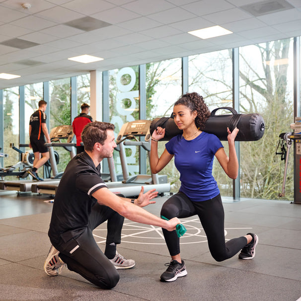 Een sporter doet in de oefenzaal een oefening met een gewicht op haar schouders. Een sportfysiotherapeut begeleidt de oefening en Wil je na een blessure of operatie snel en goed revalideren? Dat kan bij Anna TopSupport. Bijvoorbeeld na een operatie aan je voorste kruisband of je meniscus.