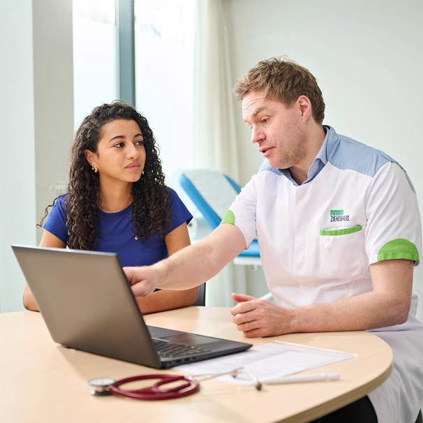 Een patiënt zit aan tafel met de dokter die uitleg geeft en iets aanwijst op een laptop. Het Basisplus Sport Medisch Onderzoek is geschikt als je zonder klachten sport en tussen de 12 en 35 jaar oud bent.