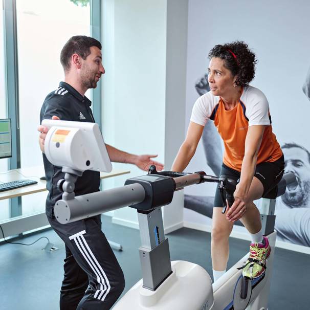 Een sporter zit op een hometrainer. De inspanningsfysioloog geeft de sporter uitleg. Bij bikefit passen we jouw fiets aan op jouw lichaam. Dit helpt om je fietsprestaties te verbeteren en blessures te voorkomen.