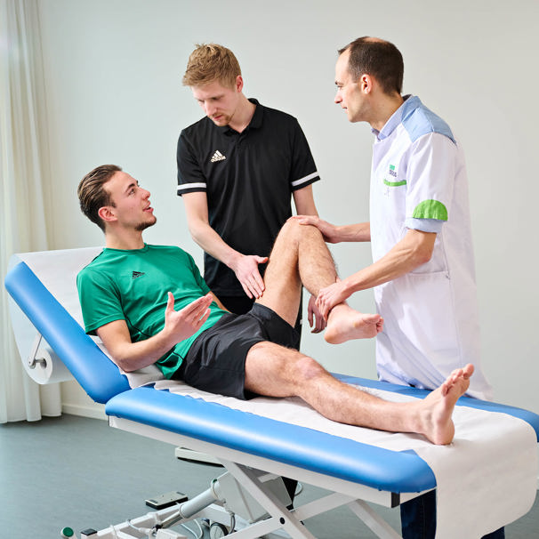 Een patiënt zit op een onderzoeksbed. De dokter en sportfysiotherapeut onderzoeken de heup van de patiënt. Anna TopSupport onderzoekt de oorzaak van heupblessures of liesblessures zoals een slijmbeursontsteking.